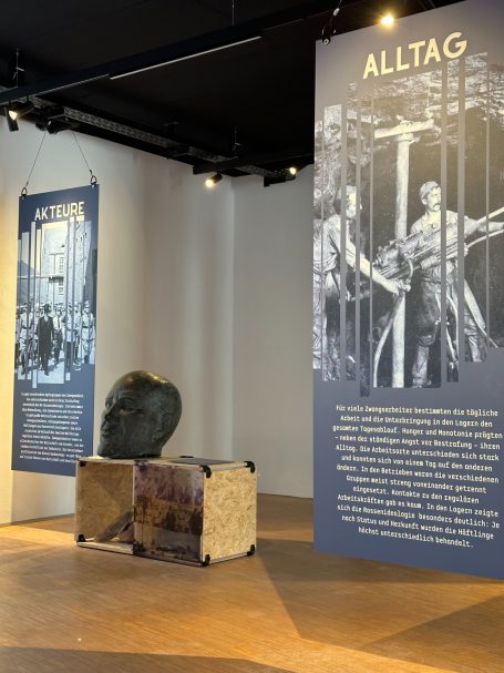 Drei große Wandtafeln in einem Ausstellungsraum mit einer Skulptur im Vordergrund.